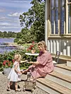 En kvinna i rödvit rutig klänning sitter på en trappa vid ett sommarhus och räcker en sak till ett litet barn.