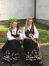 To unge kvinner i østfoldbunad sitter på en benk med blomster i fanget foran en hvit vegg.