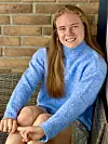 Ung kvinne i blå genser sitter og smiler foran en murvegg.