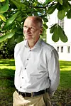 Mann i lys skjorte står under grønne blader foran en bygning.
