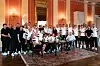Stor gruppe poserer i en elegant sal med røde vegger, lysekroner og stort teppe.