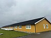 Lang gul bygning med svart tak ved en grønn gresslette under grå himmel.