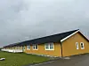 Lang gul bygning med svart tak ved en grønn gresslette under grå himmel.