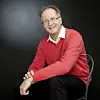 Smilende mann i rød genser sitter på stol foran mørk bakgrunn.
