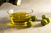 Olivenolje helles i en liten glassbolle med grønne oliven ved siden av.