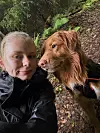 Nær selfie av en person og en brun hund i en skogssti med grønn vegetasjon.