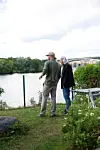 Två personer står vid ett staket vid vatten med grönska, molnig himmel och en bro i bakgrunden.