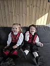 To små barn i bunad sitter på en grå sofa foran en trevegg.