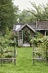 Liten trädgårdsentré med växthus, blomsterbäddar och trähus omgiven av grönska.