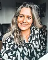 Smilende kvinne innendørs i mørk mønstret skjorte.
