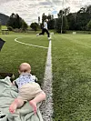 Baby ligger på et teppe ved kanten av en kunstgressbane mens en løper beveger seg forbi i bakgrunnen.
