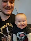 En voksen og en smilende baby sitter tett sammen innendørs, begge i svarte Guns N' Roses-klær.