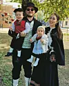 En familie i tradisjonelle drakter poserer utendørs med to små barn foran en bygning og trær.