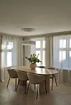 Lyst spisestueinteriør med rundt bord, fire stoler, store vinduer og en vase med blomster.