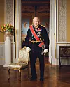 En mann i mørk seremoniuniform står i et elegant, klassisk rom ved siden av en polstret stol.