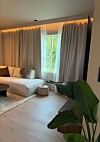 Lyst soverom med seng, store gardiner, vindu og grønne planter i forgrunnen.