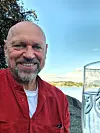 Smilende mann i rød skjorte sitter ved vannkanten med et glass i forgrunnen.