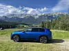 Blå SUV parkert på en gresslette foran skogkledde fjell under blå himmel.