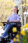 Mari Storstein sitter i en elektrisk rullestol på en sti, med gule høsttrær og blomster i bakgrunnen.