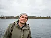 Mann i grønn jakke og grå lue står ved vannet på en overskyet dag.
