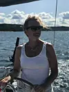 Trude i solbriller og hvit topp står ved roret på en seilbåt, med sjø og skogkledd kyst bak.