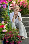 Forfatter Trude Teige sitter på steintrapper i en hage, omgitt av rosa og røde blomster.