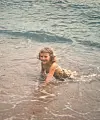Et barn bader i grunt sjøvann nær en sandstrand, med små bølger rundt kroppen.