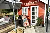 Trude Teige sitter på en treterrasse med en kopp, foran et rødt hus med hvite vinduskarmer.