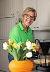 Wenche i grønn skjorte lager kaffe på et lyst kjøkken, bak en vase med gule, hvite og oransje tulipaner.