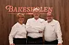 Tre personer i hvite kokkejakker står foran en trevegg med skiltet «Bakeskolen».