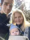 To voksne står tett med en baby i bæresjal utendørs, med sollys og trær i bakgrunnen.
